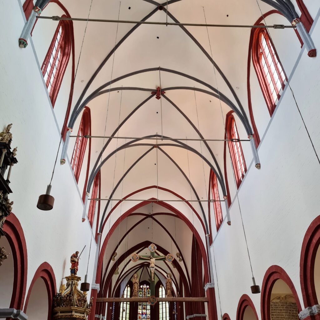 Das gotische Deckengewölbe. Es ist weiß. Die Fenster haben rötliche Umrandungen und die Bögen schwarze.