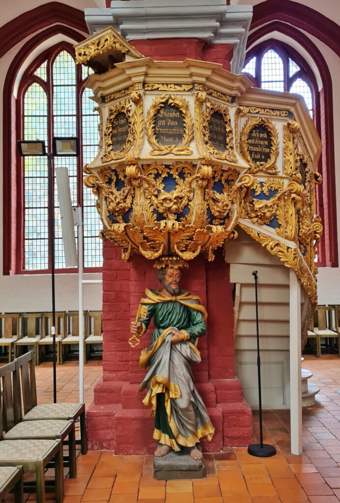 Die Kanzel, die en einem roten Kirchenpfeiler angebracht ist. Sie ist mit viel Gold verziert. Außerdem sind auf runden Platten Bibelzitate. Unter der Kanzel steht eine männliche Statue, so das es aussieht, als ob sie die Kanzel auif dem Kopf trägt. Der bärtige Mann ist barfuss. Er trägt ein weites dunkelgrünes Gewand mit goldenen Borten. Mit der linken Hand rafft er es nach oben, so dass die graue Unterseite des Gewandes sichtbar wird. In der anderen Hand hält er einen großen goldenen Schlüssel.