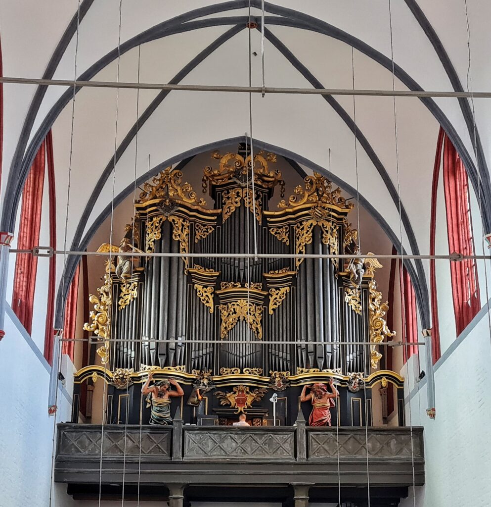 Eine große dunkle Orgel mit goldenen Verzierungen. Man sieht den Oberkörper des Organisten von hinten. Links und rechts davon stehen zwei große männliche Statuen in weiten Gewänder (links grün, rechts rot). Sie haben die Hände abgewinkelt erhoben, so das es aussieht als ob sie die Orgel tragen.