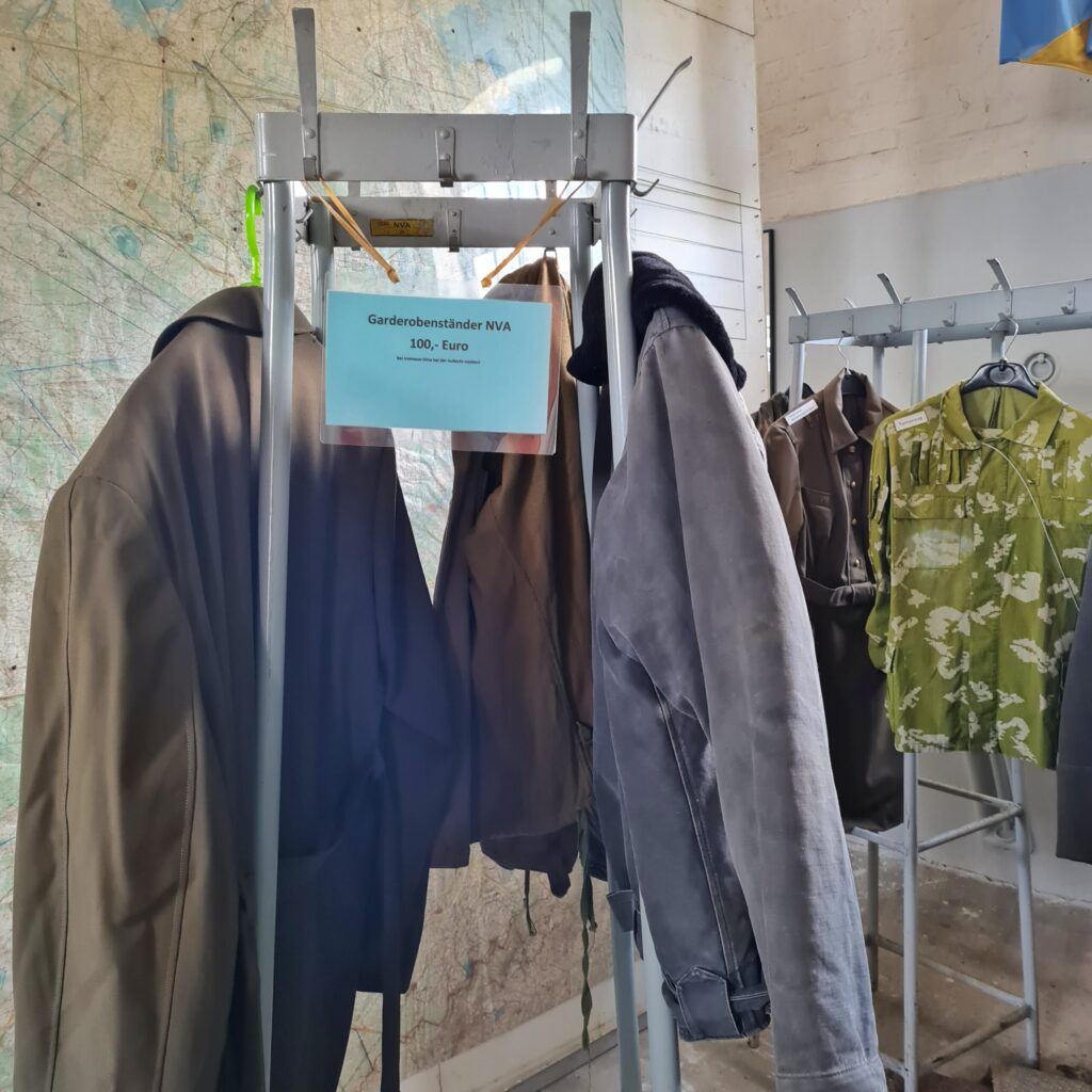 Ein viereckiger Metallständer, an dem mehrere Jacken hängen. Außerdem ein Schild: Garderobenständer NVA 100 €. Im Hintergrund ein länglicher Garderobenständer, an dem ein Mantel und ein grün-weiß-gemustertes Hemd hängt.
