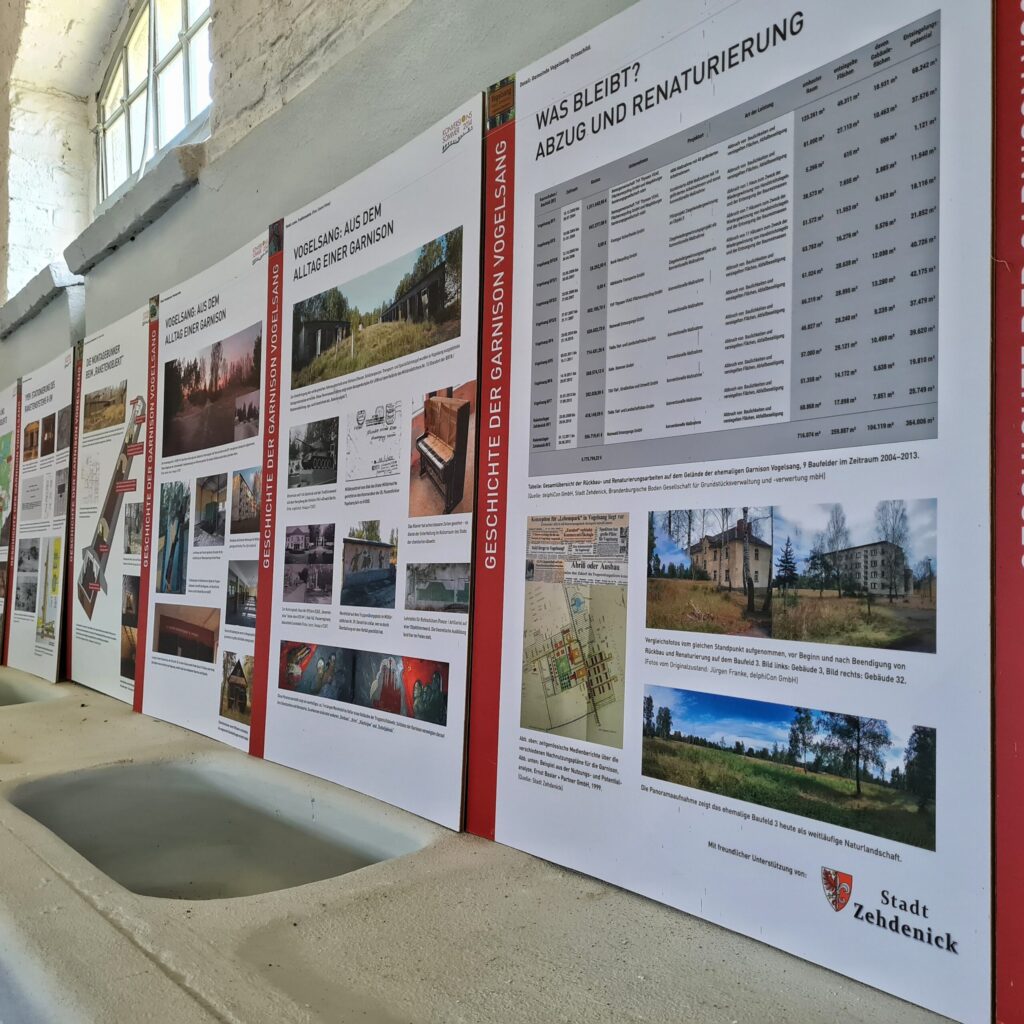 Ausstellungstafeln an einer hellen Wand, seitlich fotografiert. Darauf viele Fotos und Erklärungen. Auf den Tafeln, die am dichtesten an der Kamera sind, stehen die Überschriften "Vogelsang: Aus dem Alltag einer Garnison" und "Was bleibt? Abzug und Renaturierung".