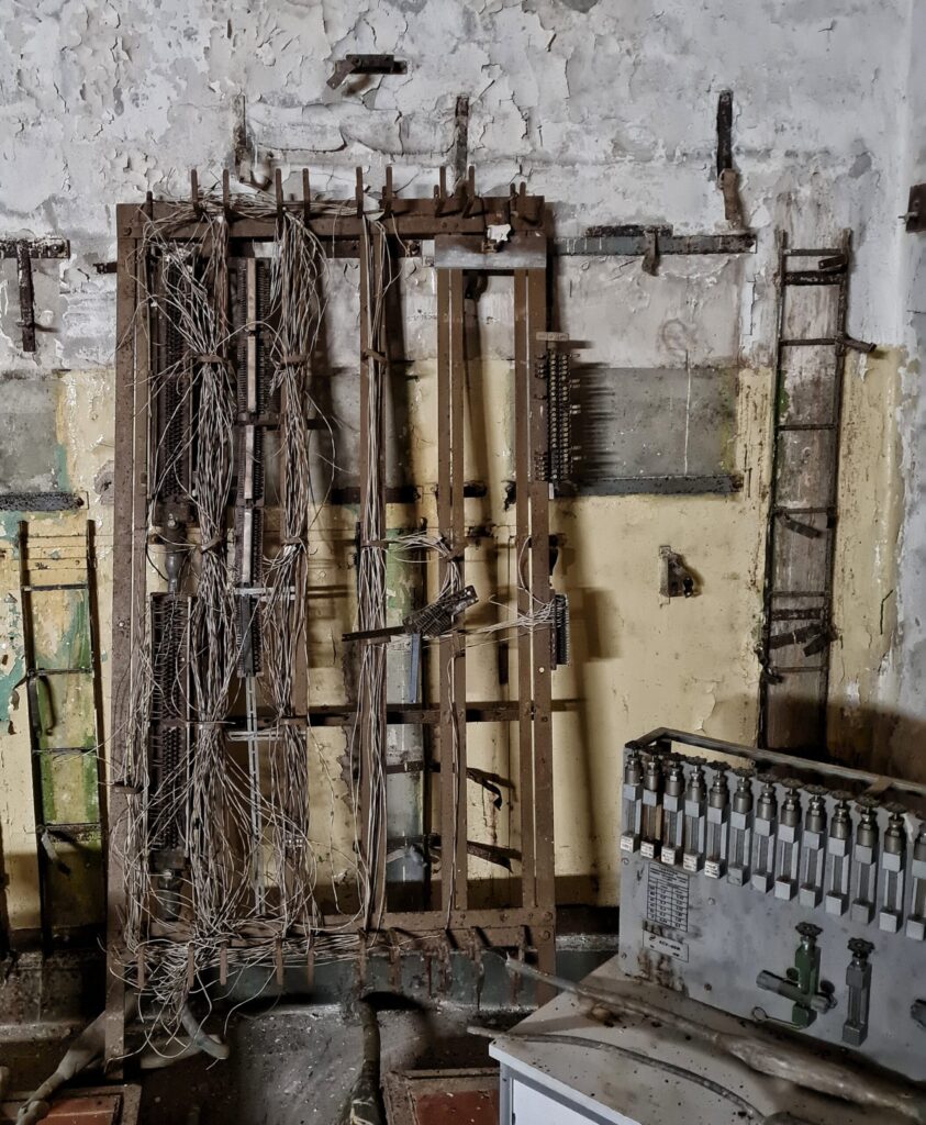 An eine Mauer gelehnt ein Metallgitter mit senkrechten Streben. Daran hängen zusammengerollt Kabel herunter. Rechts ein Gerät mit Schaltern.