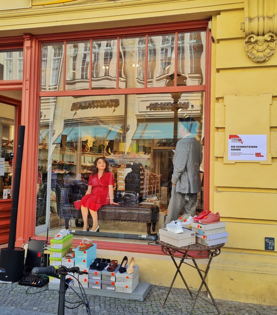 Ein Schuhladen in einem gelben Gebäude mit Verzierungen. Das Schaufenster ist von rötlich angestrichenem Holz umrandet. Oben sind Sprossen. Vor dem Schaufenster steht ein kleiner Tisch mit Schuhen darauf und danben auf dem Boden Schuhkartons, auf denen Schuhe stehen. Daneben eine Lautsprecheranlage. Im Schaufenster stitzt eine Schuapoelerin im roten Kleid auf einem alten Ledersofa. Ein Schauspielerin einem hellgrauen Anzug steht seitlich von ihr. Die beiden reden miteinander. An der Wand neben dem Schaufenster hängt ein Schild, auf dem steht: Schauspielfenster. Die schmutzigen Hände. Von Jean-Paul Sartre. mit Guido Lambrecht und Mascha Schneider.Ab 14. Oktober 2022. Hans Otto Theater.