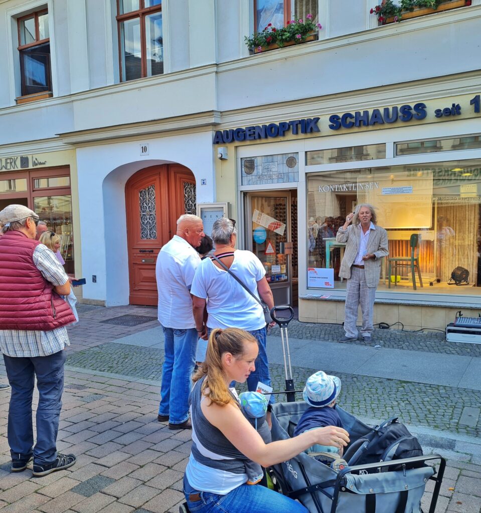Ein Augenoptikerladen. Im Schaufenster ein Holztisch, ein Holzstuhl und eine helle Tafel. Auf dem -Schaufenaster steht: Kontaktlinsen, und unten ist ein Schild angebracht, auf dem "Kinder der Sonne" steht. Vor dem Schaufenster ein Schauspieler im hellen Anzug, der redet und gestikuliert. Im Vordergrund mehrere Menschen, weitgehend mit dem Rücken zur Kamera, die zuschauen.