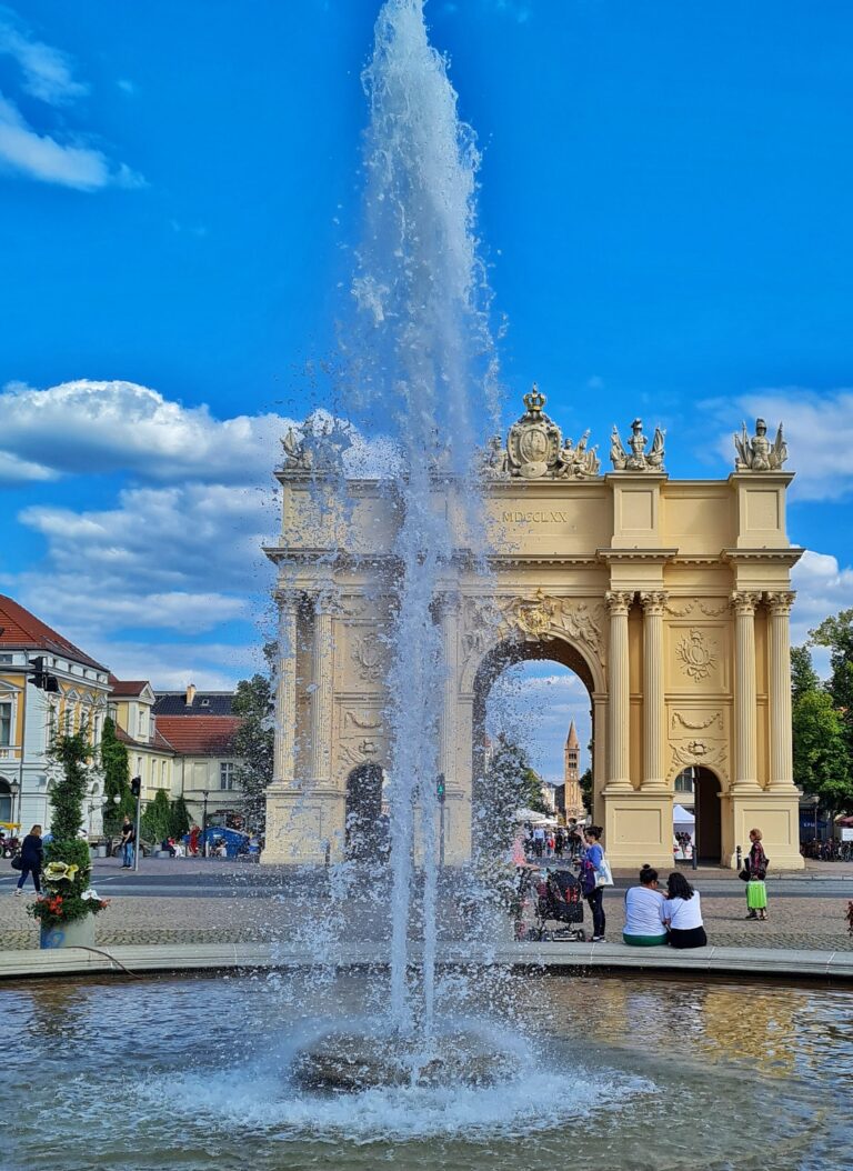 Das Brandenburger Tor in Potsdam fotogerofaiert duch die Wasserfontäne des Springbrunnes auf dem Luisenplatz. Auf der Umrandung des Springbrunnes sitzen einige Menschen.