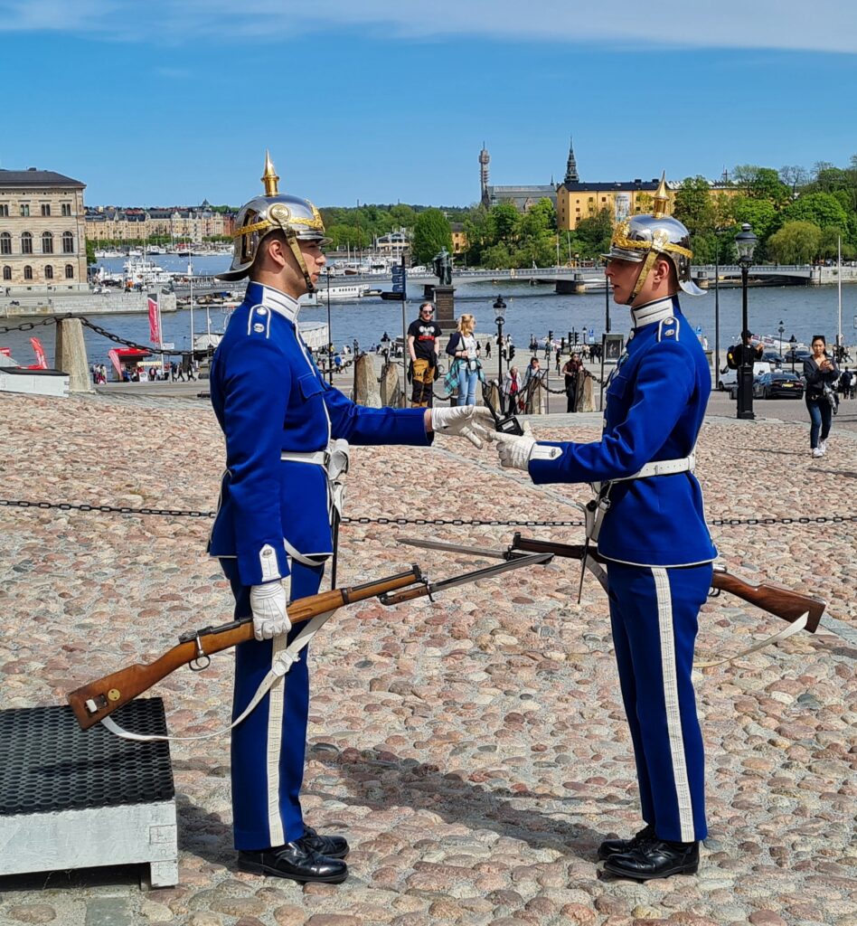 Das Bild zeigt zwei junge Wachsoldaten in blauen Uniformen mit silbernen Helmen mit goldenen Spitzen und mit Gewehren auf einem Kopfsteinpflasterplatz in einer Stadt. Sie stehen sich gegenüber und ihre Gewehre sind vor ihnen gekreuzt. Sie werden von mehreren Menschen im Hintergrund beobachtet. Dahinter ist eine Wasserfläche mit einer niedrigen Brücke und Booten zu sehen, gesäumt von aneinander stehenden großen, alten Gebäuden und Bäumen. 