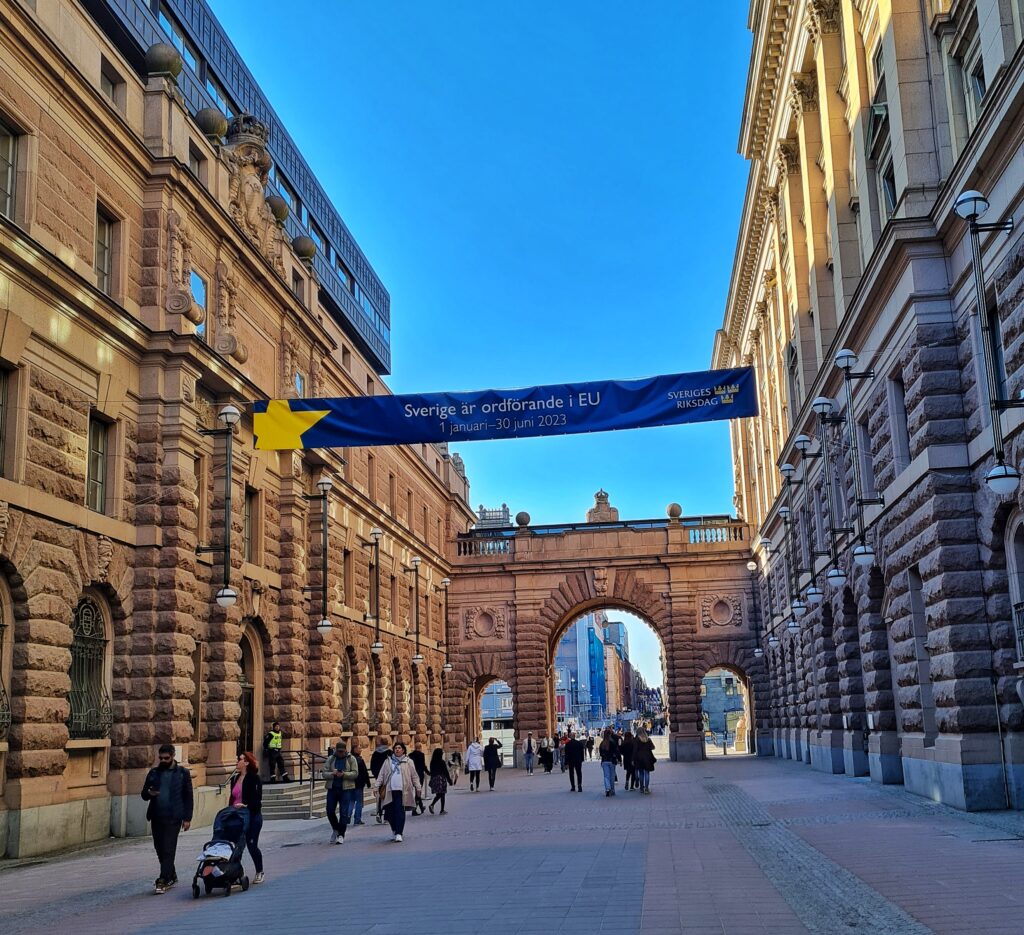 Ein breiter Durchgang mit alten, prächtigen Gebäuden auf beiden Seiten und einem großen Torbogen im Hintergrund. Mehrere Spaziergänger laufen zwischen den Gebäuden hindurch. Darüber hängt ein blaues Banner mit der Aufschrift “Sverige är ordförande i EU” (Schweden ist der Vorsitzende der EU). Der Banner hat einen gelben Stern darauf. Der Himmel ist blau und das Wetter ist klar. 