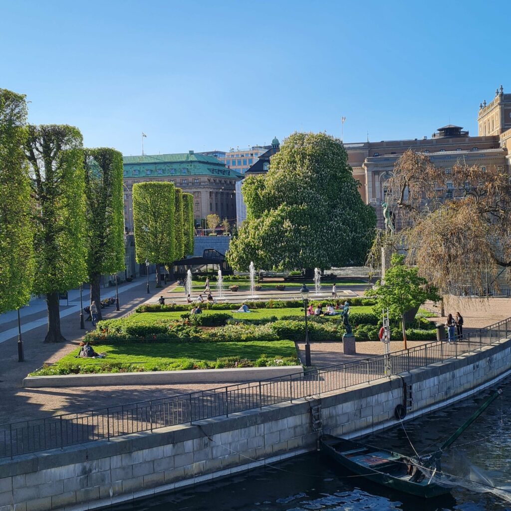 Ein Park mit  Rasenflächen, Blumen, niedrigen Büschen, Bäumen, einem Springbrunnen mit mehreren kleinen Fontänen und einer Statue. Eine Reihe von Bäumen auf der rechten Seite sind viereckig beschnitten. Mehrere Menschen laufen auf den Wegen oder sitzen auf dem Rasen. Im Hintergrund sind große, alte Gebäude zu sehen. Rechts vorne ist eine Wasserfläche mit einer Kaimauer zu sehen. Dort ist ein Ruderboot angebunden.