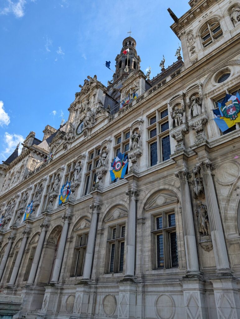Detailaufnahme der Fassade des Hôtel de Ville in Paris, mit Säulen, Statuen und Fahnen vor blauem Himmel.